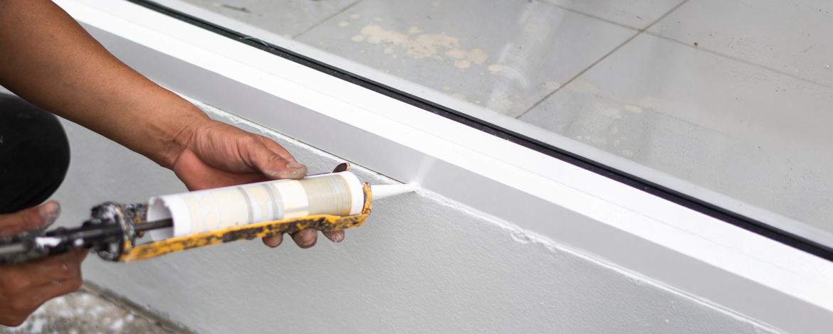 Person sealing a gap along a window frame with a caulking gun to prevent air leaks in a house