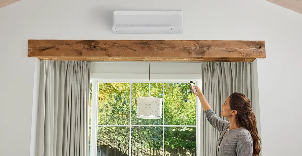 A person using a remote control to operate a wall-mounted mini-split air conditioning unit in a bright living room with large windows and curtains