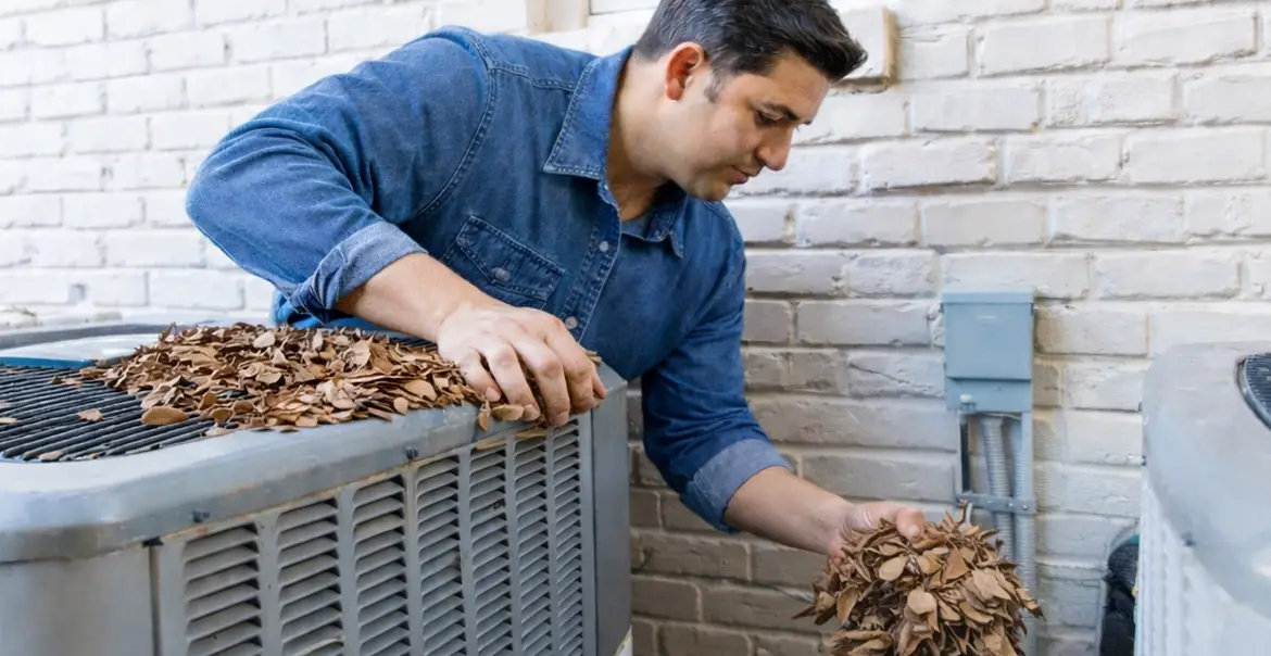 Person clearing dried leaves off an outdoor heat pump unit next to a brick wall, showing maintenance of debris around the system
