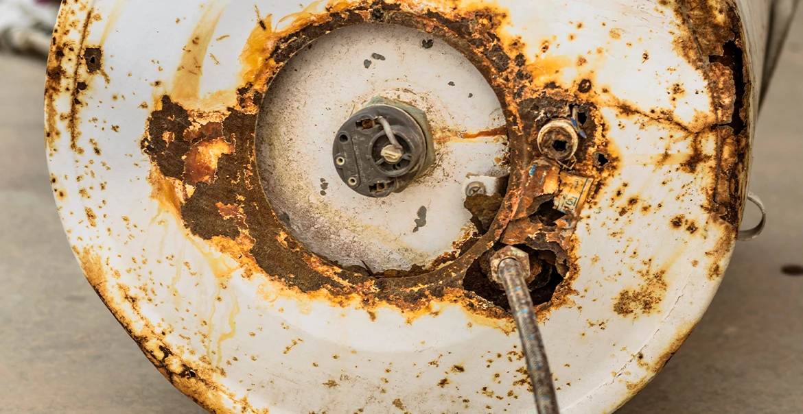 Bottom of a severely rusted and corroded water heater showing extensive metal deterioration and damaged components