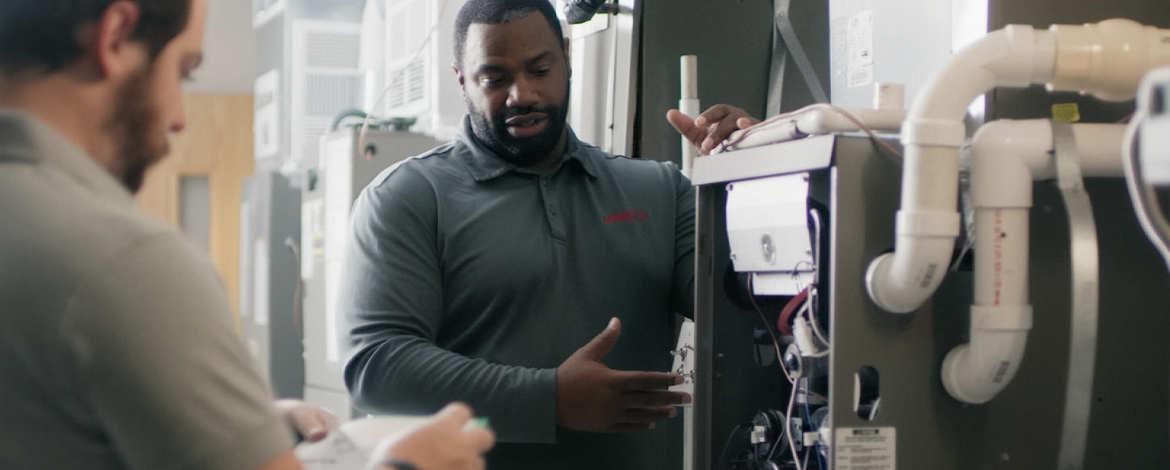Technician inspecting and explaining the components of an HVAC furnace system, with visible pipes and internal parts of the unit